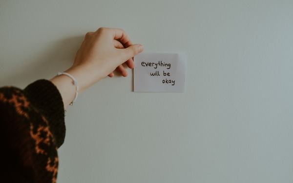 Eine Hand hält einen Zettel an die Wand, auf dem steht: „Everything will be okay“.