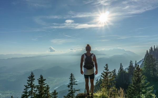 Eine Wanderin steht auf einem sonnigen Berggipfel und blickt über eine weite Landschaft.